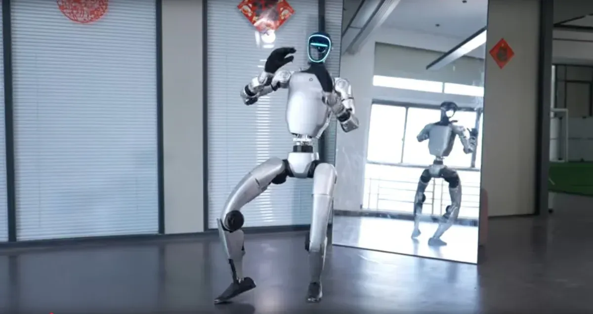 Bipedal robot with teal torso, black limbs, and silver lower legs stands against a cloudy sky.