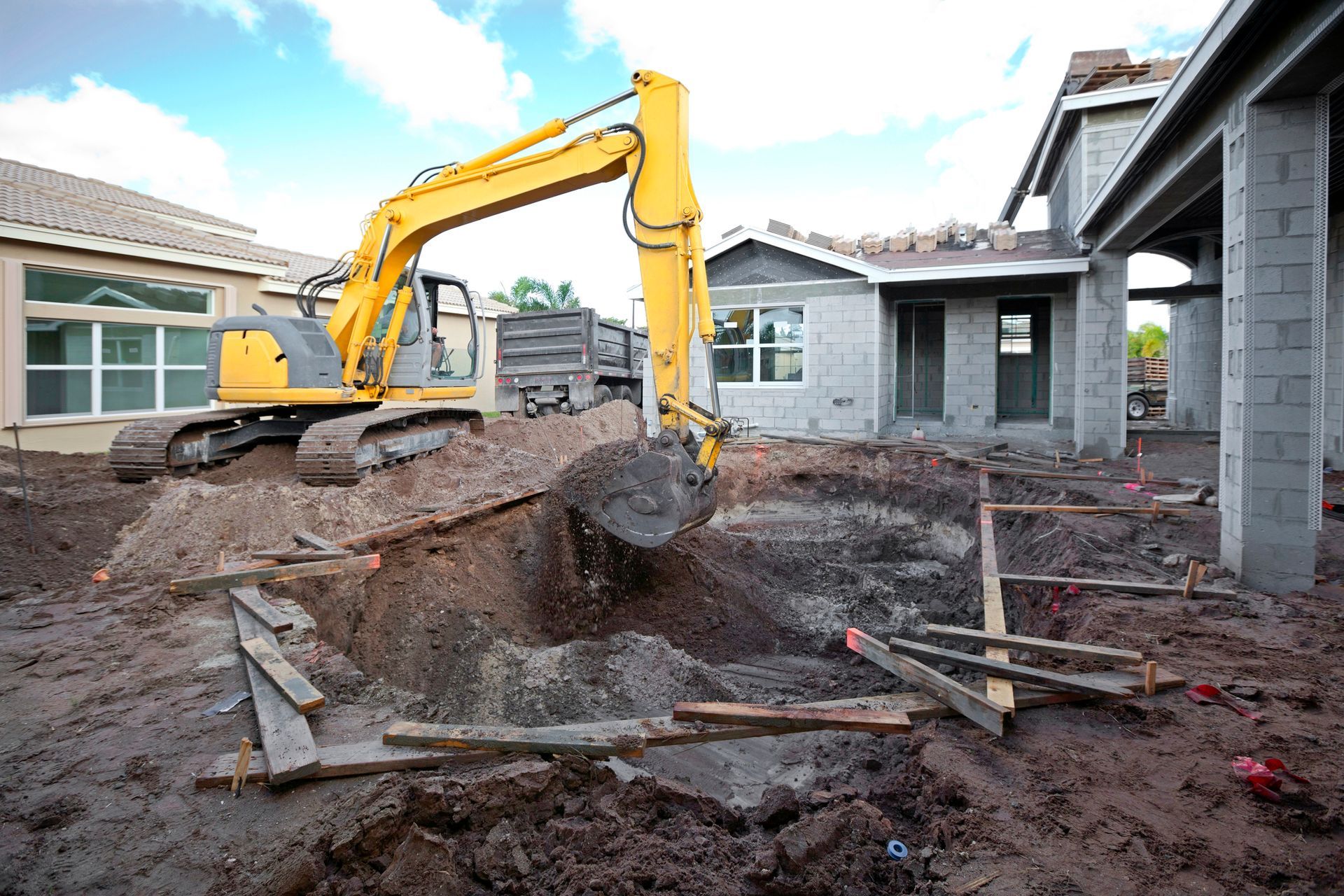 A yellow excavator is digging a hole in front of a house under construction.