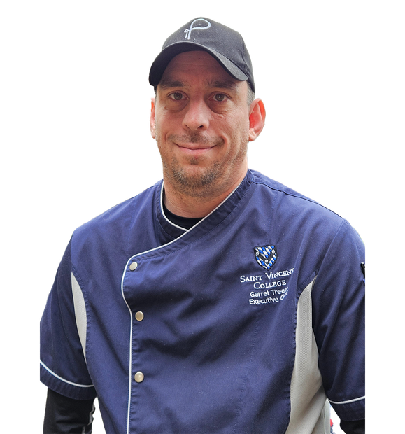 A person wearing a dark blue chef's coat with an emblem and a black baseball cap.