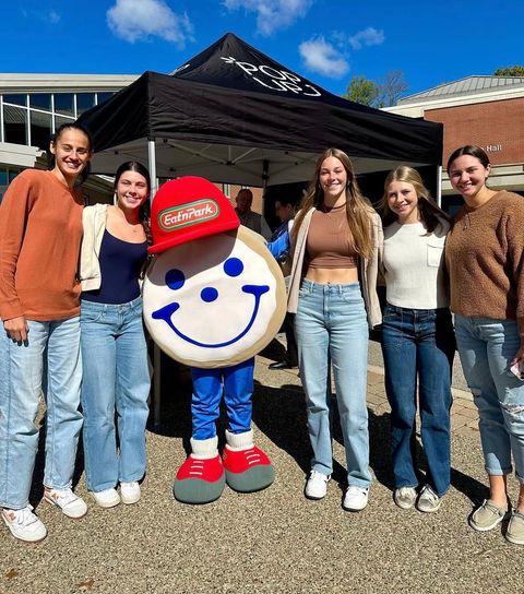 Six people pose smiling outdoors under a black canopy with a large circular cookie mascot.