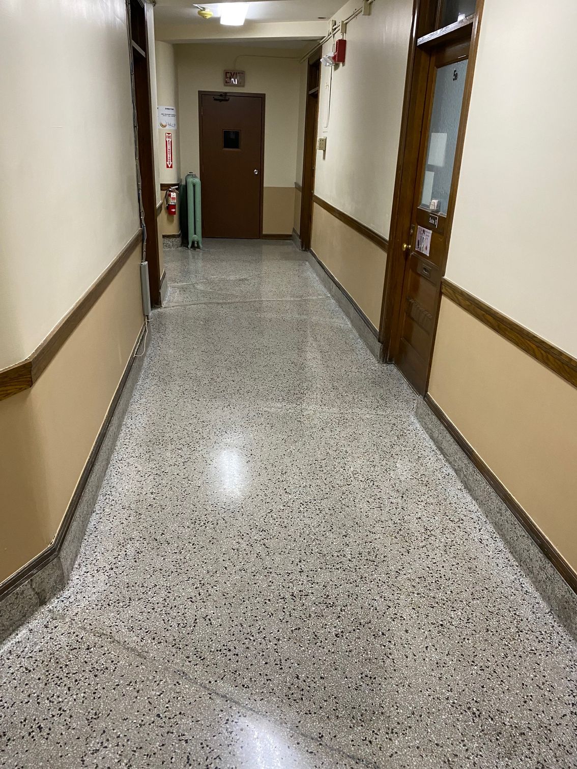 Empty hallway with beige walls, wooden doors, and speckled tile floor leading to a closed door.