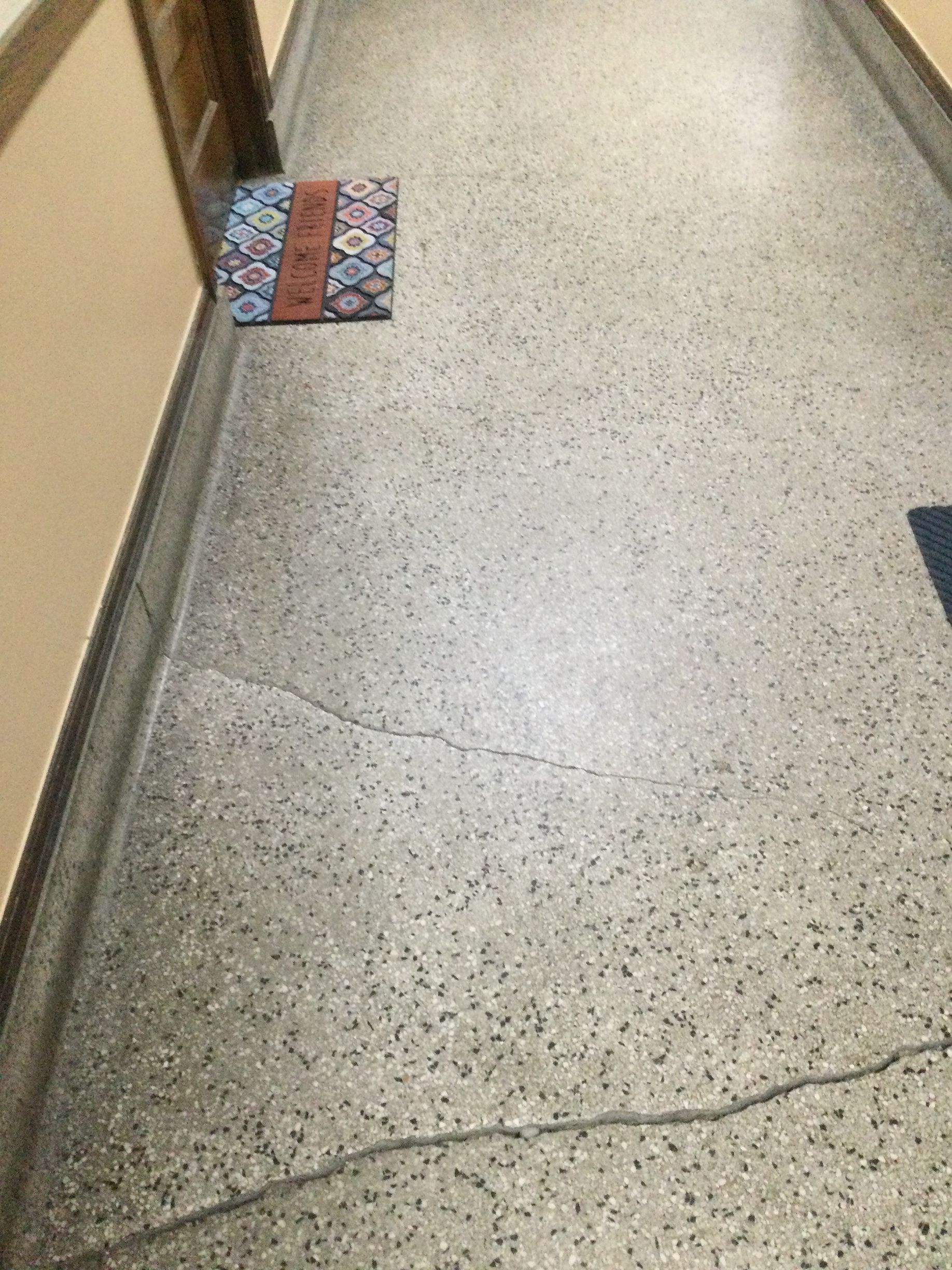 Cracked speckled hallway floor beside a wall and patterned mat near a doorway