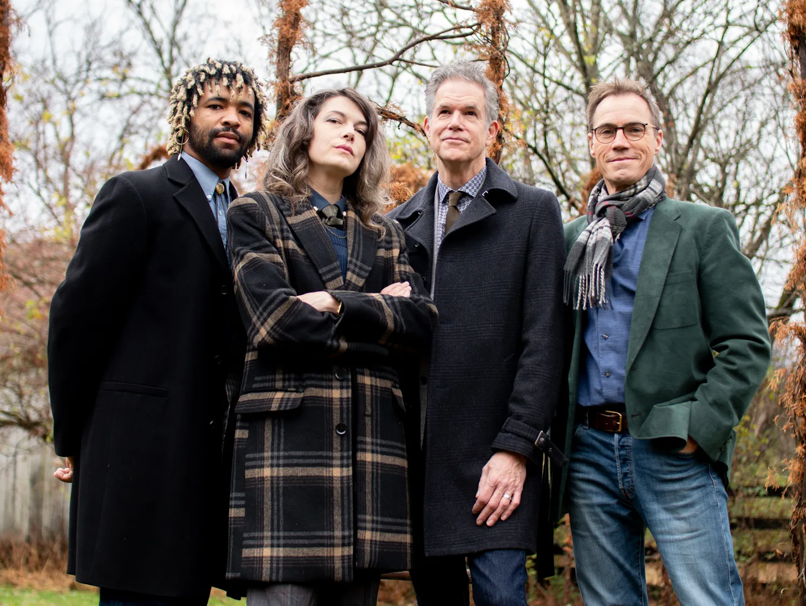 Four people standing outdoors in coats, posing in a leafless wooded area.