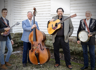 Four musicians pose outdoors with guitars, upright bass, and banjo in front of white siding.