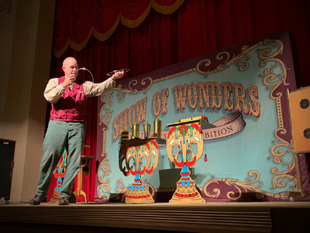Magician onstage pointing beside colorful carnival props and a “World of Wonders” backdrop