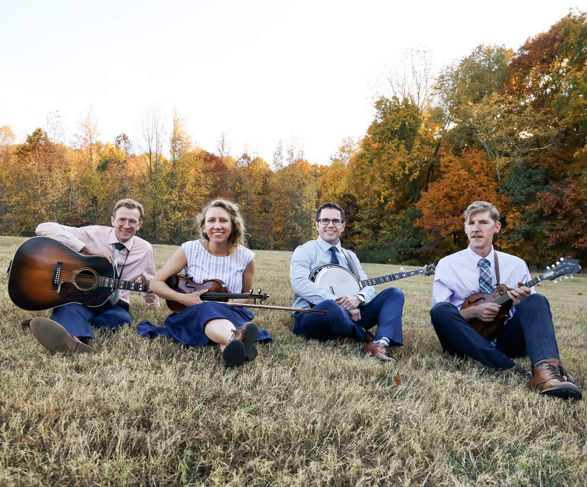 Four people sit in a grassy field with guitars, smiling in front of autumn trees.