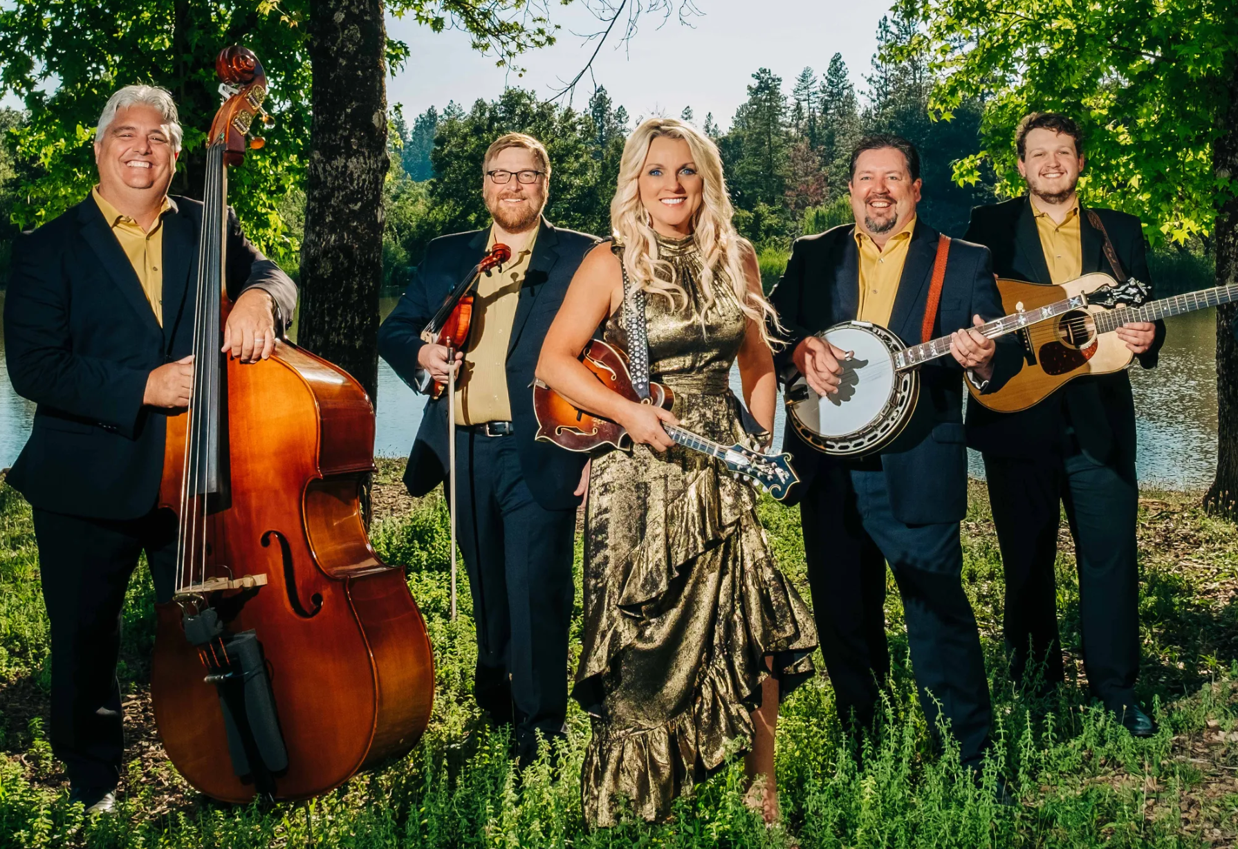 Five musicians posing outdoors in formal dark outfits, holding an upright bass, fiddle, banjo, and mandolin.