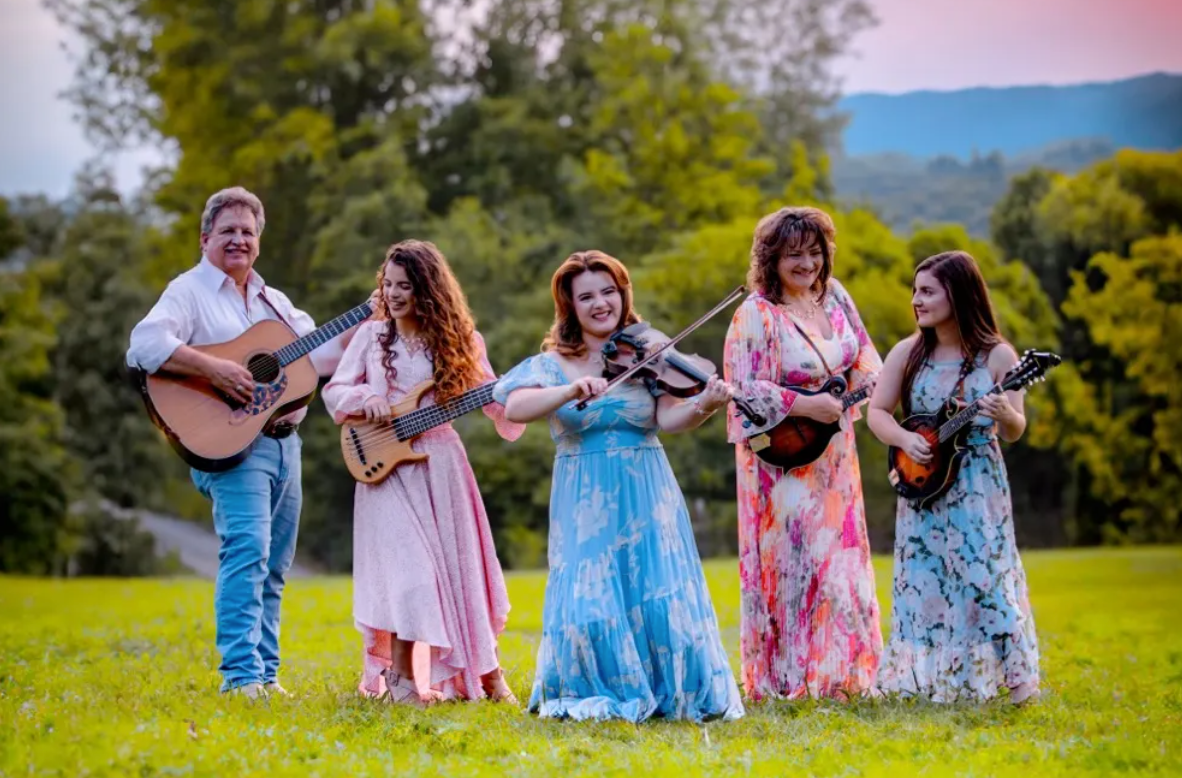 Five musicians playing string instruments in a green field with trees and mountains behind them