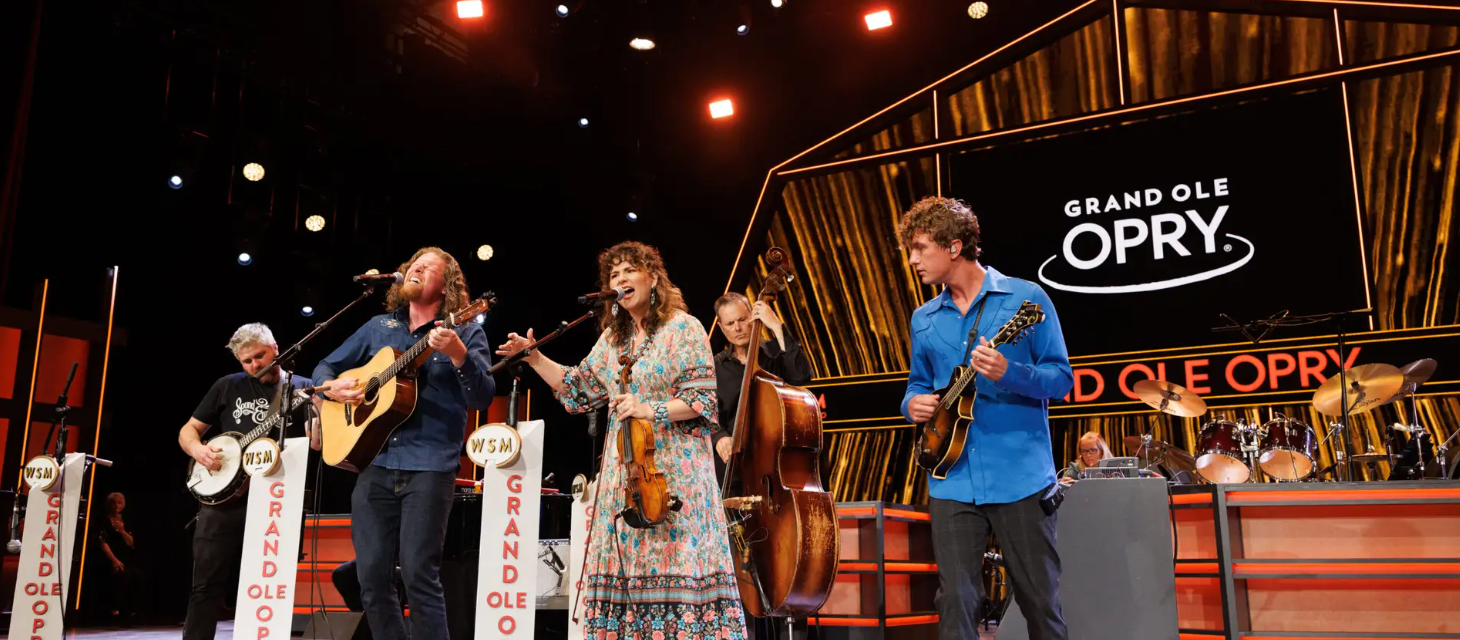 Band performing live on stage with microphones, guitars, and “Grand Ole Opry” signage in the background