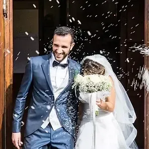 Newlyweds exiting a doorway amid confetti, groom in blue suit and bride in white dress holding bouquet