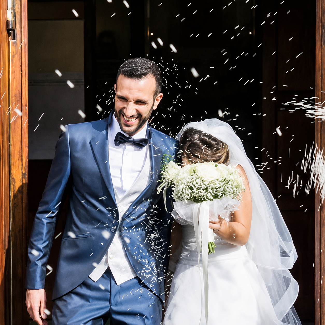 Bride and groom exiting a doorway as confetti falls, holding a bouquet and smiling