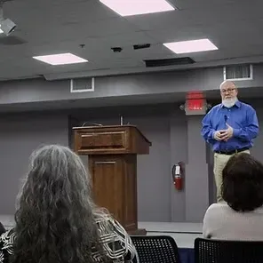 Speaker in blue shirt standing beside a podium in a meeting room, addressing an audience.