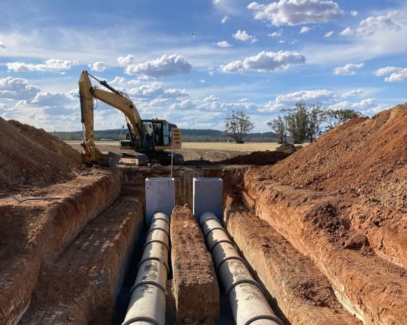 A yellow excavator is digging a trench with pipes in it.