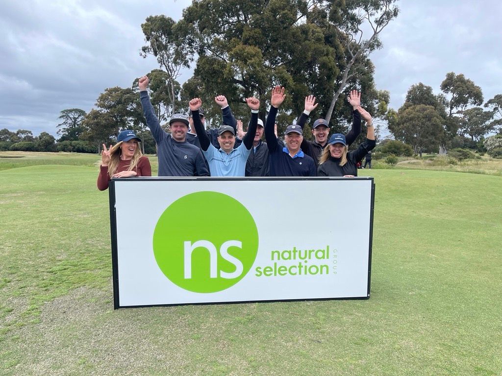 A group of people standing in a field on a golf course, cheering behind a sign for Natural Selection.