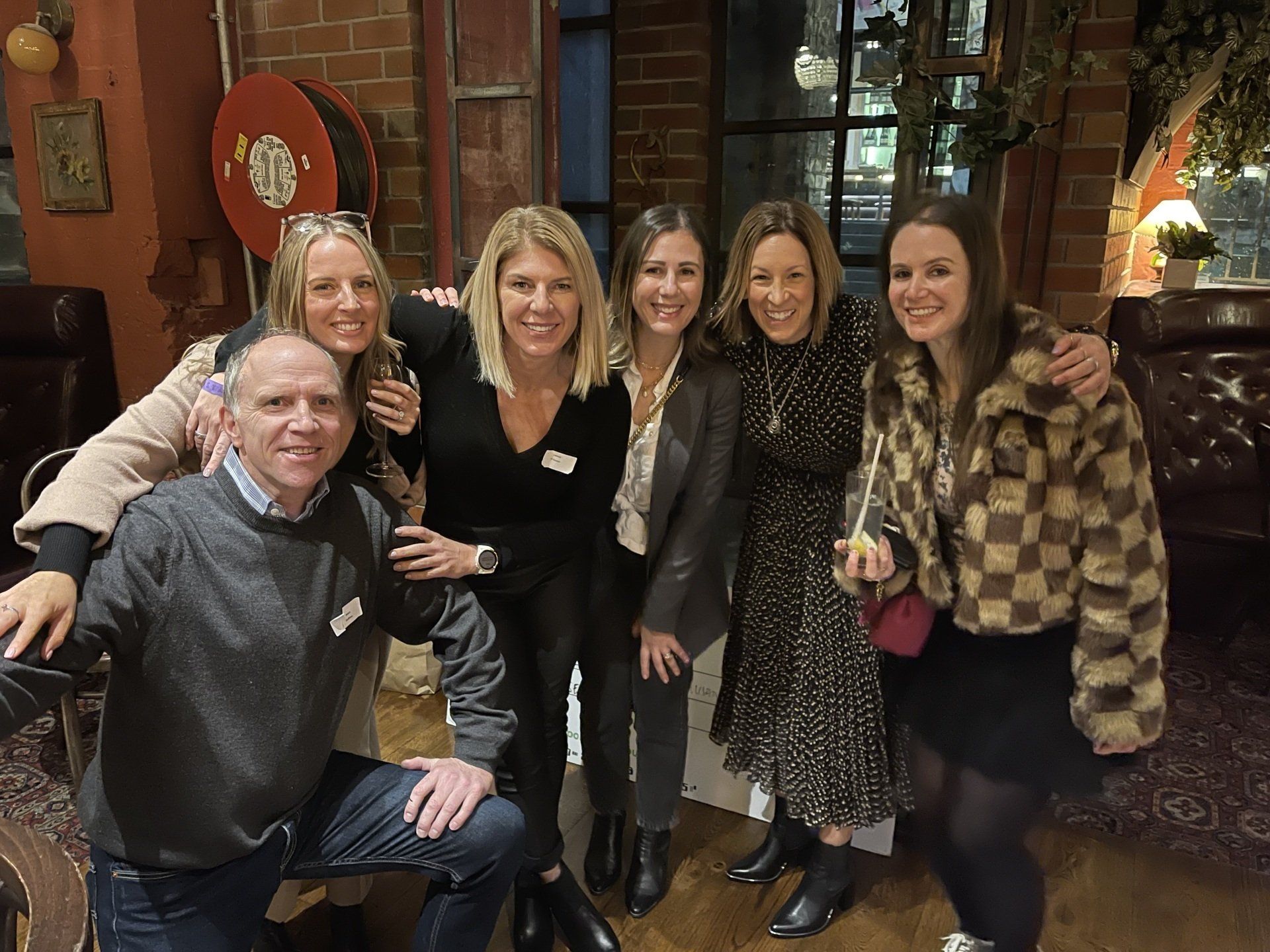 A group of six people posing for a photo in a warm-toned, dimly lit room with brick walls and casual seating.