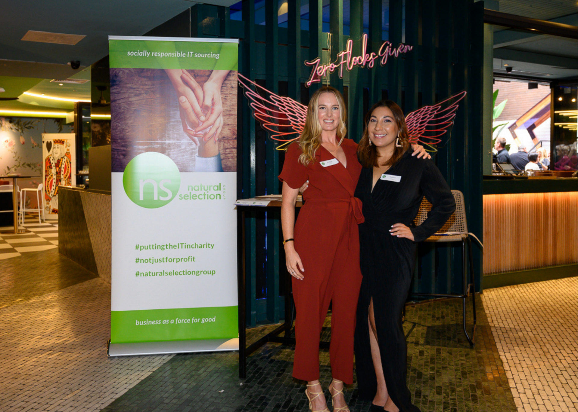 Two people stand smiling next to a branded display banner in a venue with dark decor and neon wing artwork.