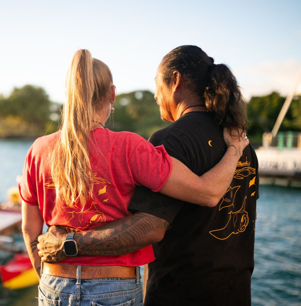 Two people with arms around each other, looking at water. One wears red, the other black, a watch on one's wrist.