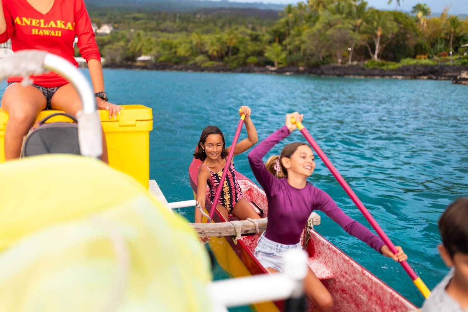 Two girls paddling a canoe in blue water, smiling, with a woman in red shirt at the back.