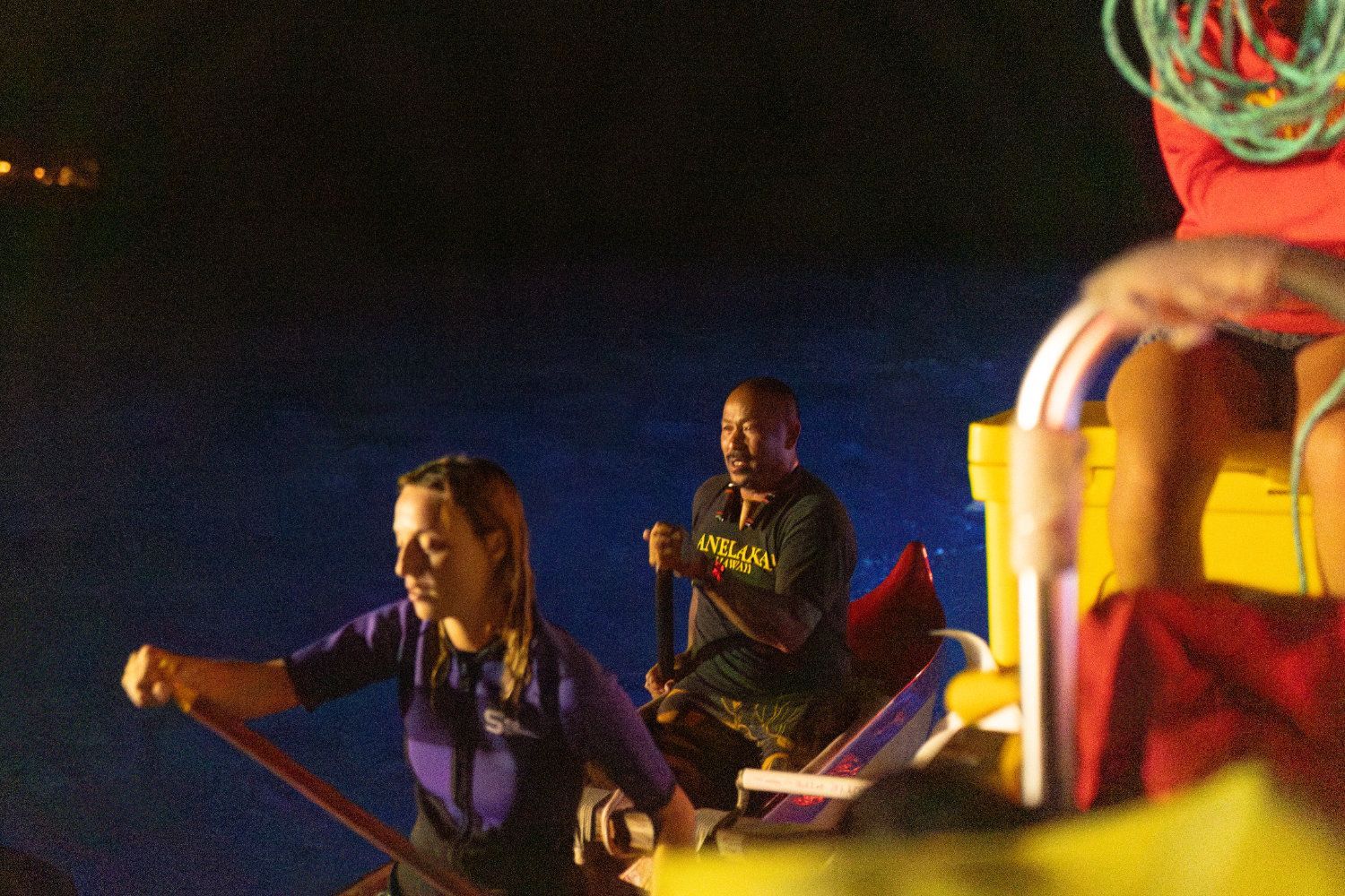 People paddling in a boat at night. Dark blue water, two people with paddles, one in purple, one in black shirt.