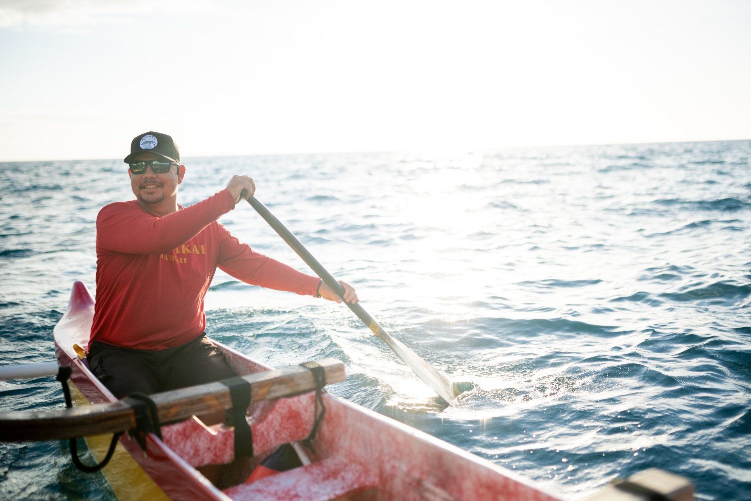Man rowing a colorful outrigger canoe on the ocean, sunlit waves in the background.