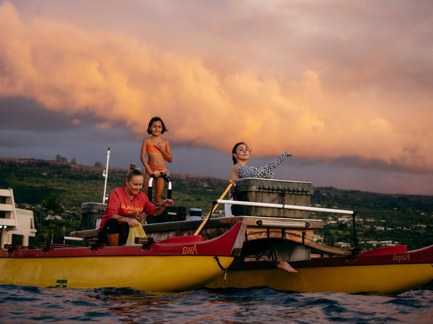 People on a yellow and red catamaran boat, enjoying sunset with pink and orange clouds.
