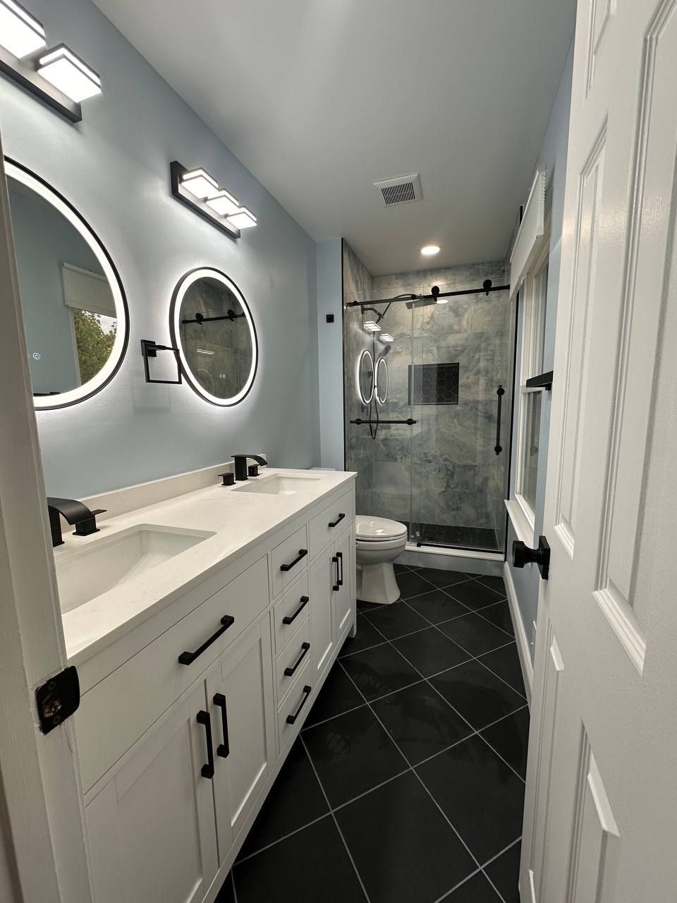 Remodeled bathroom in Elkridge, MD with white double vanity, backlit round mirrors and walk-in glass shower