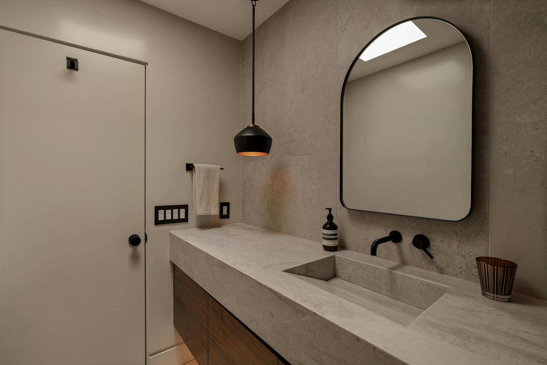 Modern bathroom vanity detail — integrated marble countertop and sink, arched black-framed mirror, matte black wall-mount faucet, black pendant light, and large format stone tile walls — Handyman On Demand, Maryland
