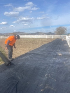 A person wearing an orange shirt unrolls a long strip of black landscape fabric on a grassy field near a white fence.