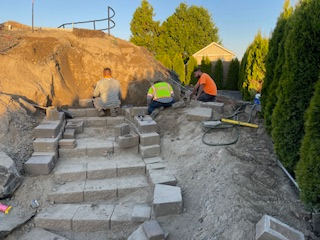 Three workers build a stone staircase into a dirt embankment outdoors during the day.