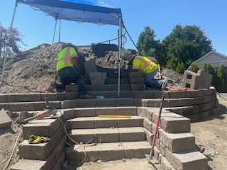 Two workers in high-visibility vests construct stone stairs on a residential hillside under a blue pop-up canopy.