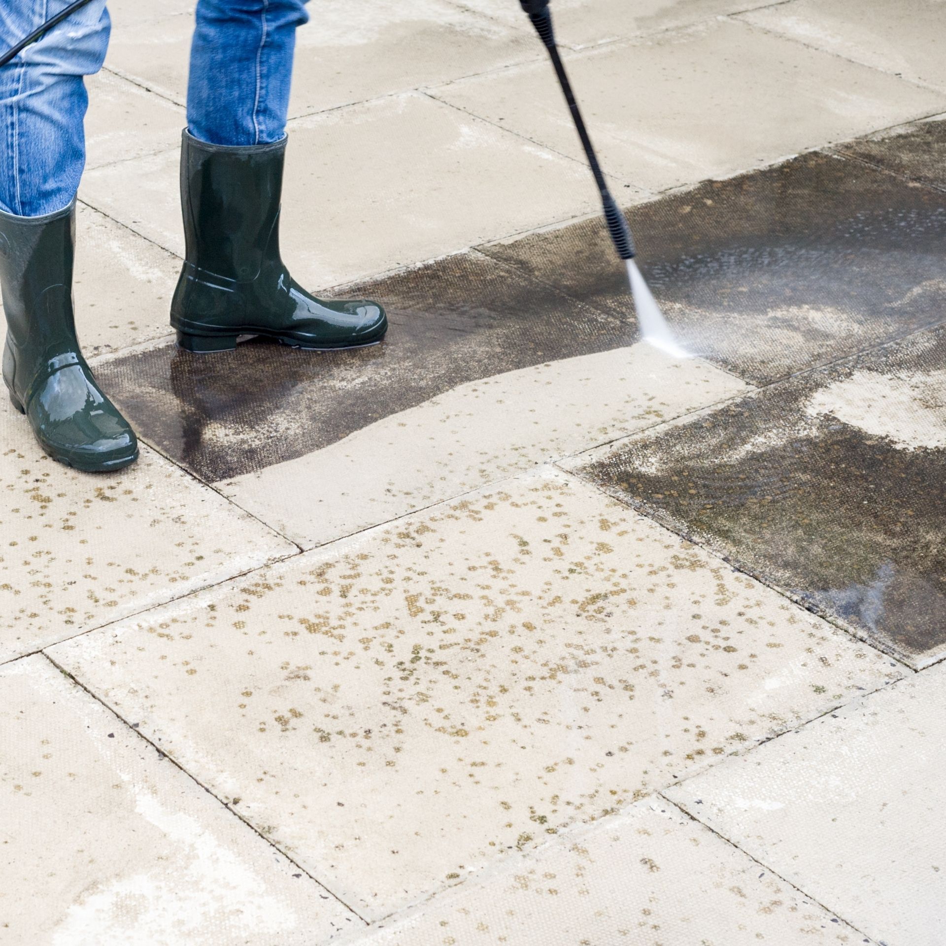 Person pressure-washing a sidewalk, with black boots and a spray wand cleaning wet concrete tiles