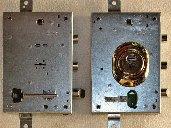 Due meccanismi di chiusura in metallo per porte, uno anteriore e uno posteriore, su sfondo bianco.