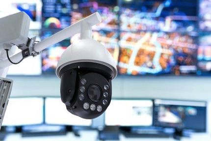Telecamera di sicurezza bianca in una sala di controllo con diversi schermi di sorveglianza sfocati sullo sfondo.