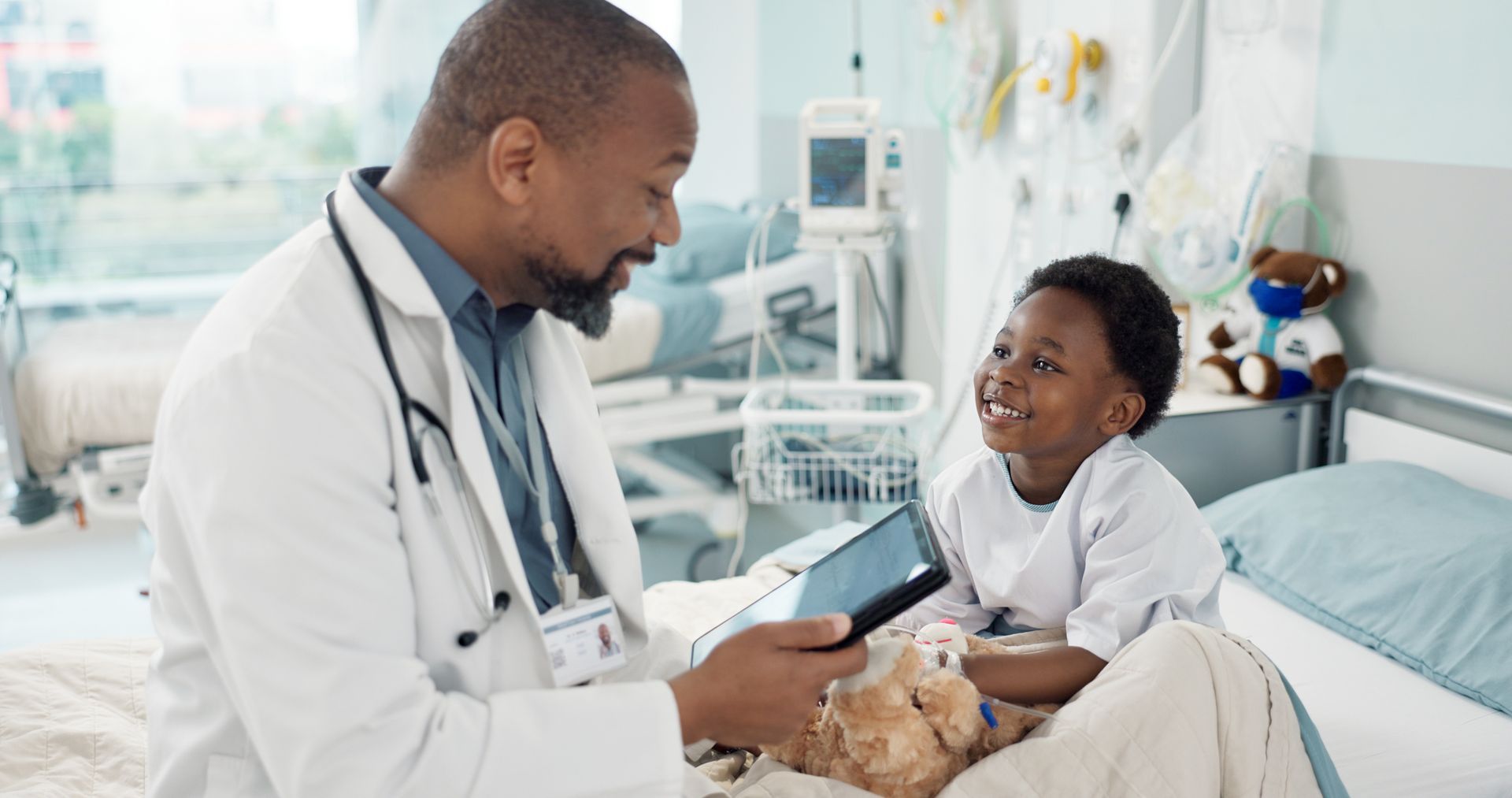 A medical professional in a white coat shows a tablet to a smiling child sitting in a hospital bed. A medical professional in a white coat shows a tablet to a smiling child sitting in a hospital bed.
