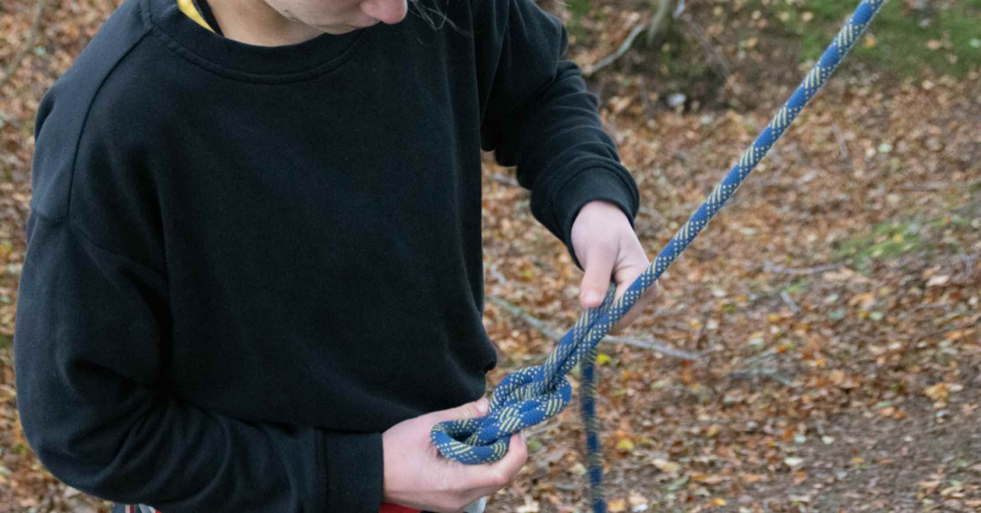 Person, der binder et blåt og hvidt reb. De har en sort sweater på, udendørs med efterårsløv.