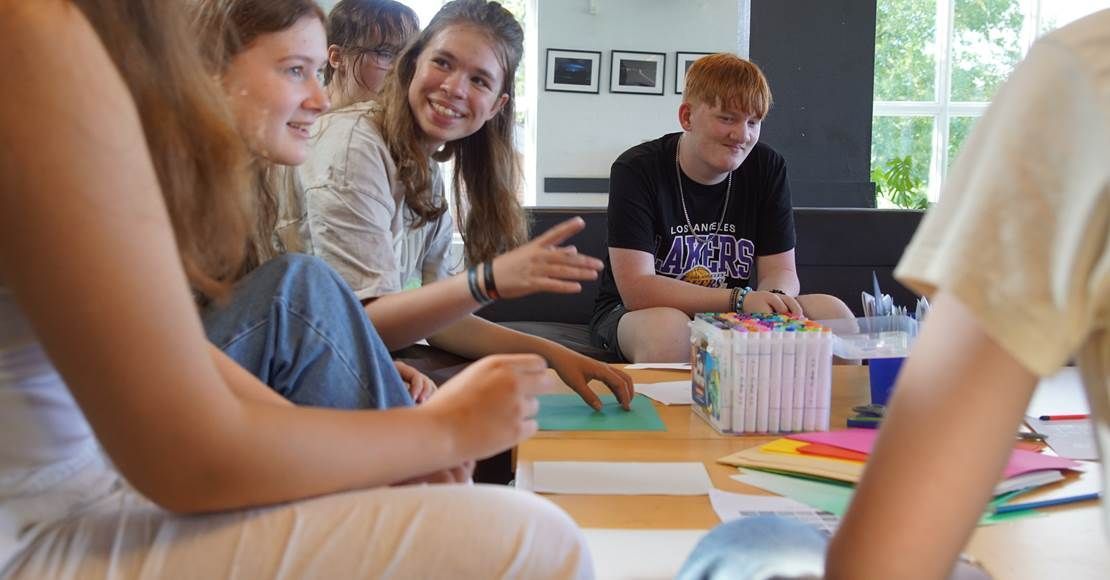Folk sidder omkring et bord, smiler, snakker og arbejder på kunstprojekter.