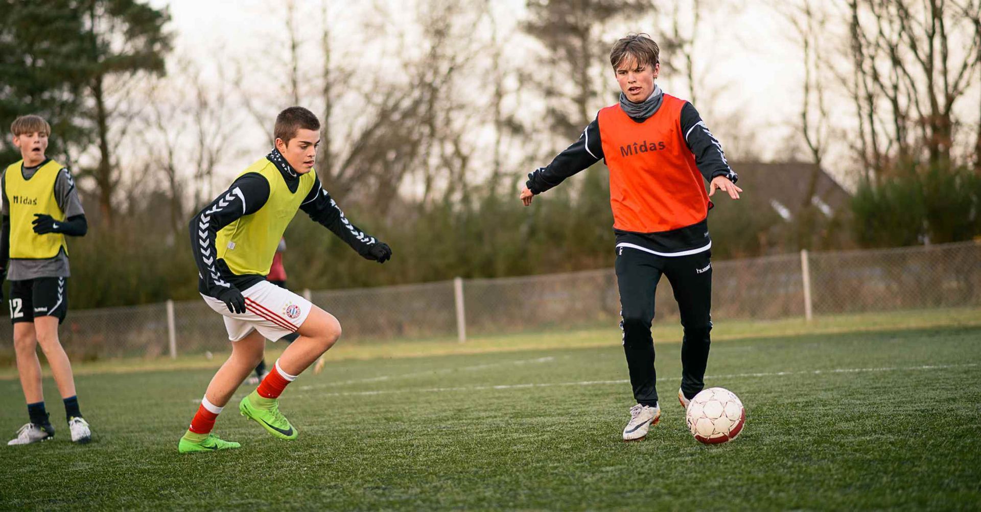 Tre fodboldspillere i veste på en grøn bane. En dribler med bolden, to andre forfølger ham.