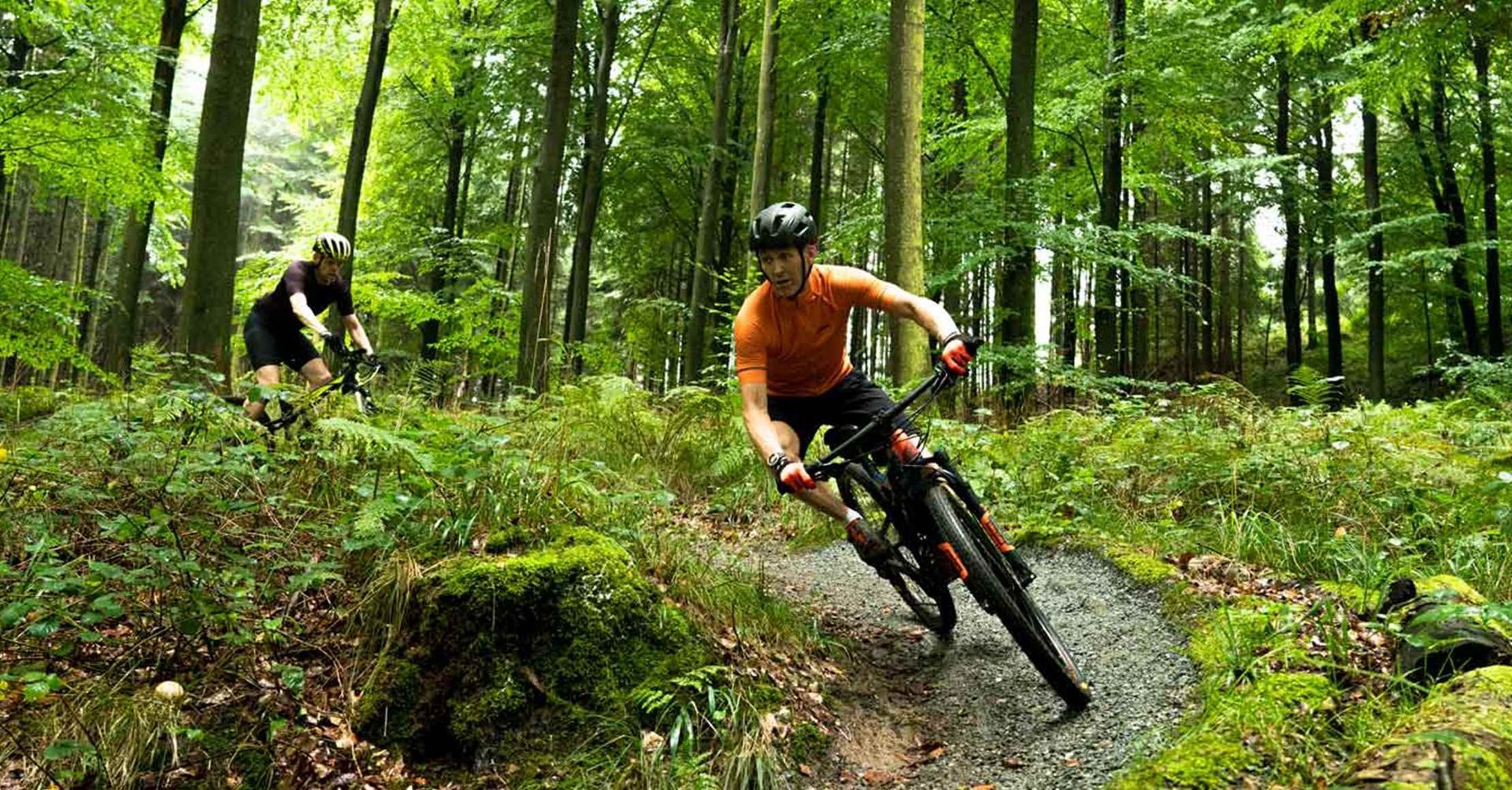 To mountainbikere på en skovsti, den ene i orange læner sig ind i et sving, de andre stier bagved.