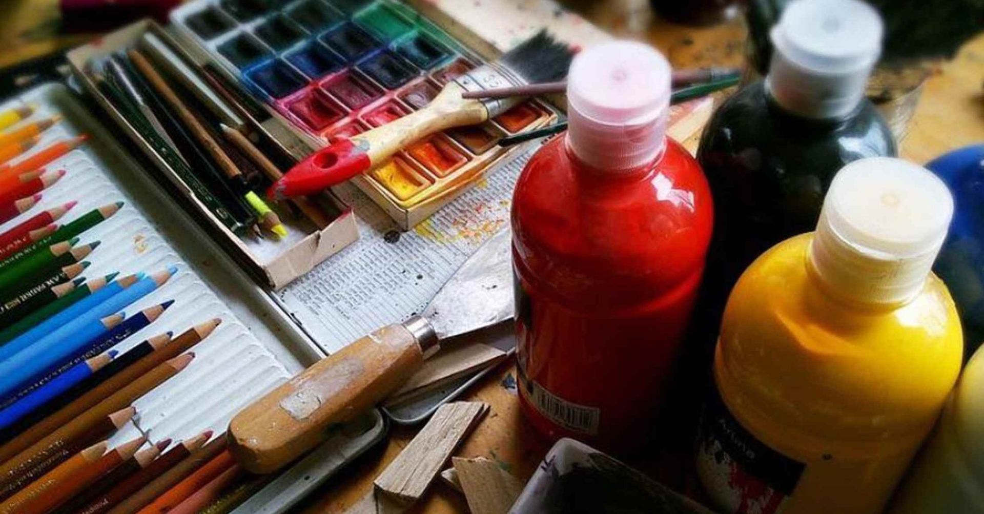 Kunstforsyninger: malingflasker, akvarelpalette, blyanter og paletknive på et bord.