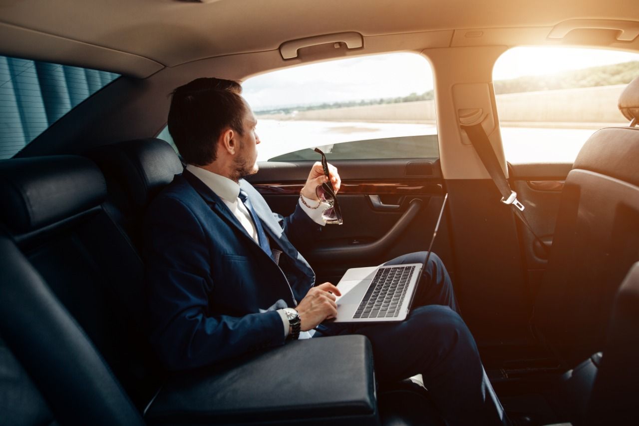Un hombre está sentado en el asiento trasero de un automóvil usando una computadora portátil y un teléfono celular.