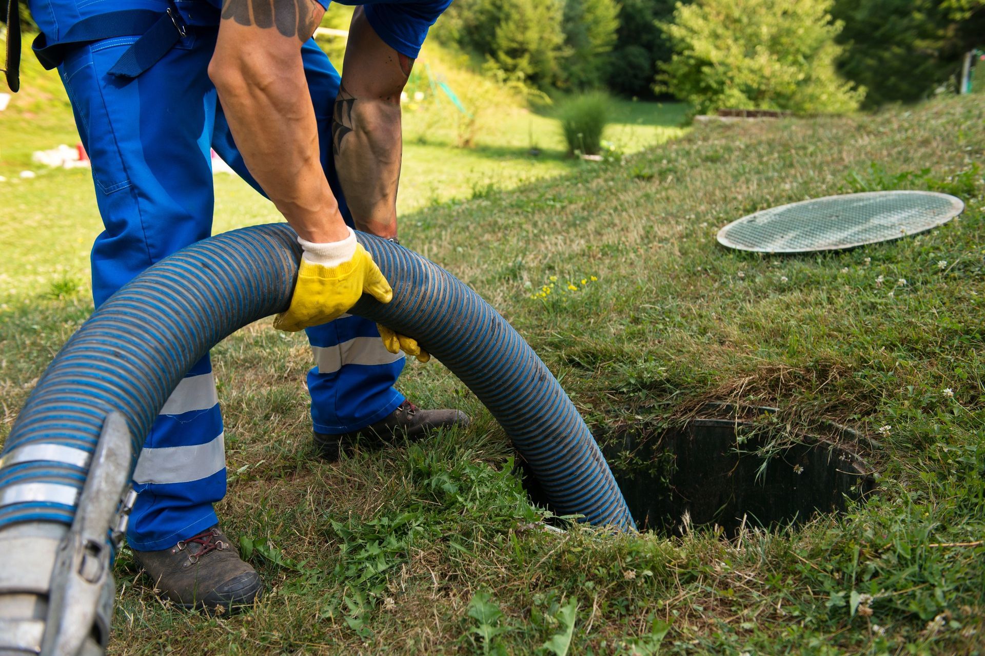 Man in blue overalls and gloves pumping out a septic tank in a grassy yard.
