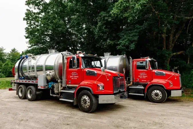 Two red tanker trucks parked on a dirt lot beside green trees