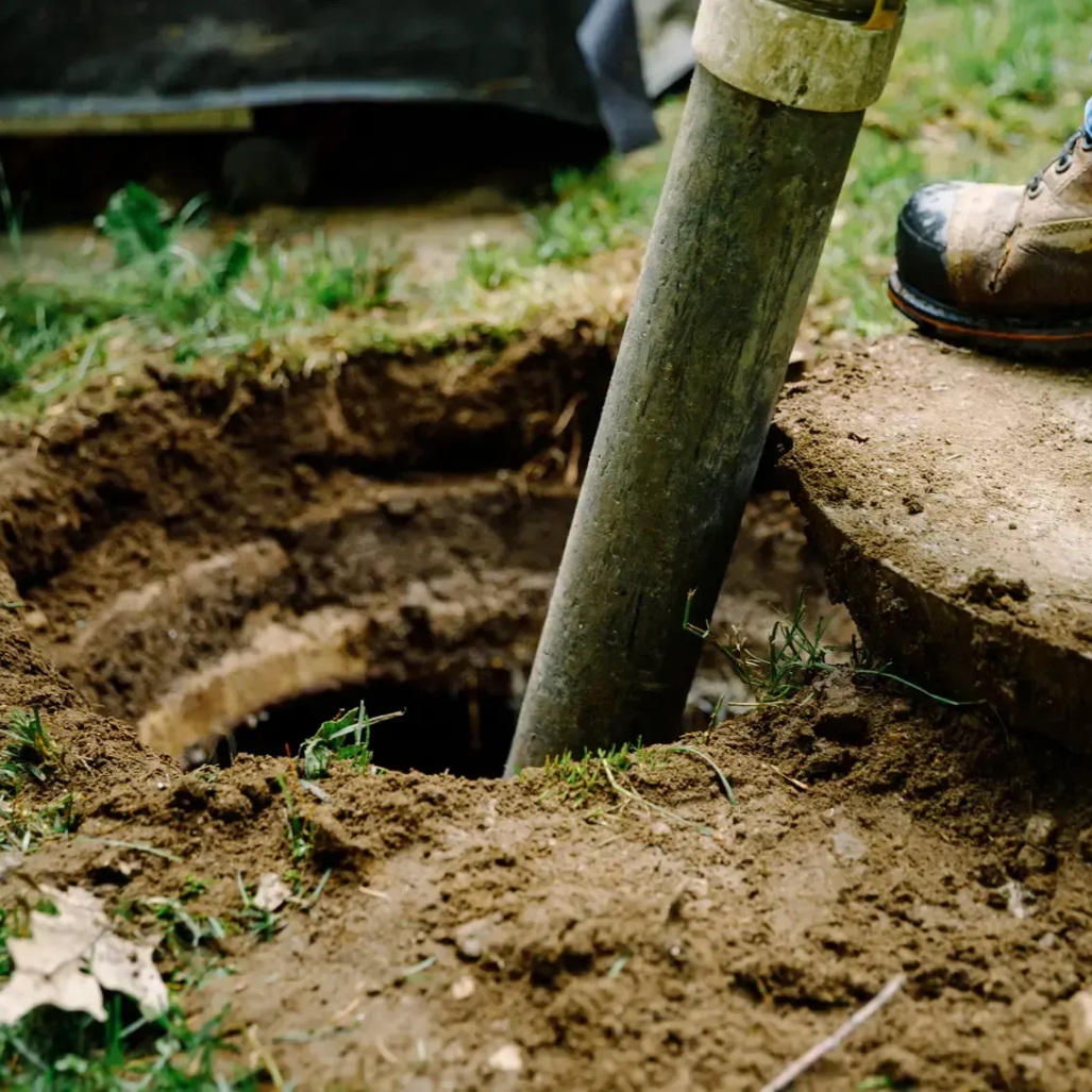 Shovel digging into a dirt hole beside a foot in a work boot outdoors