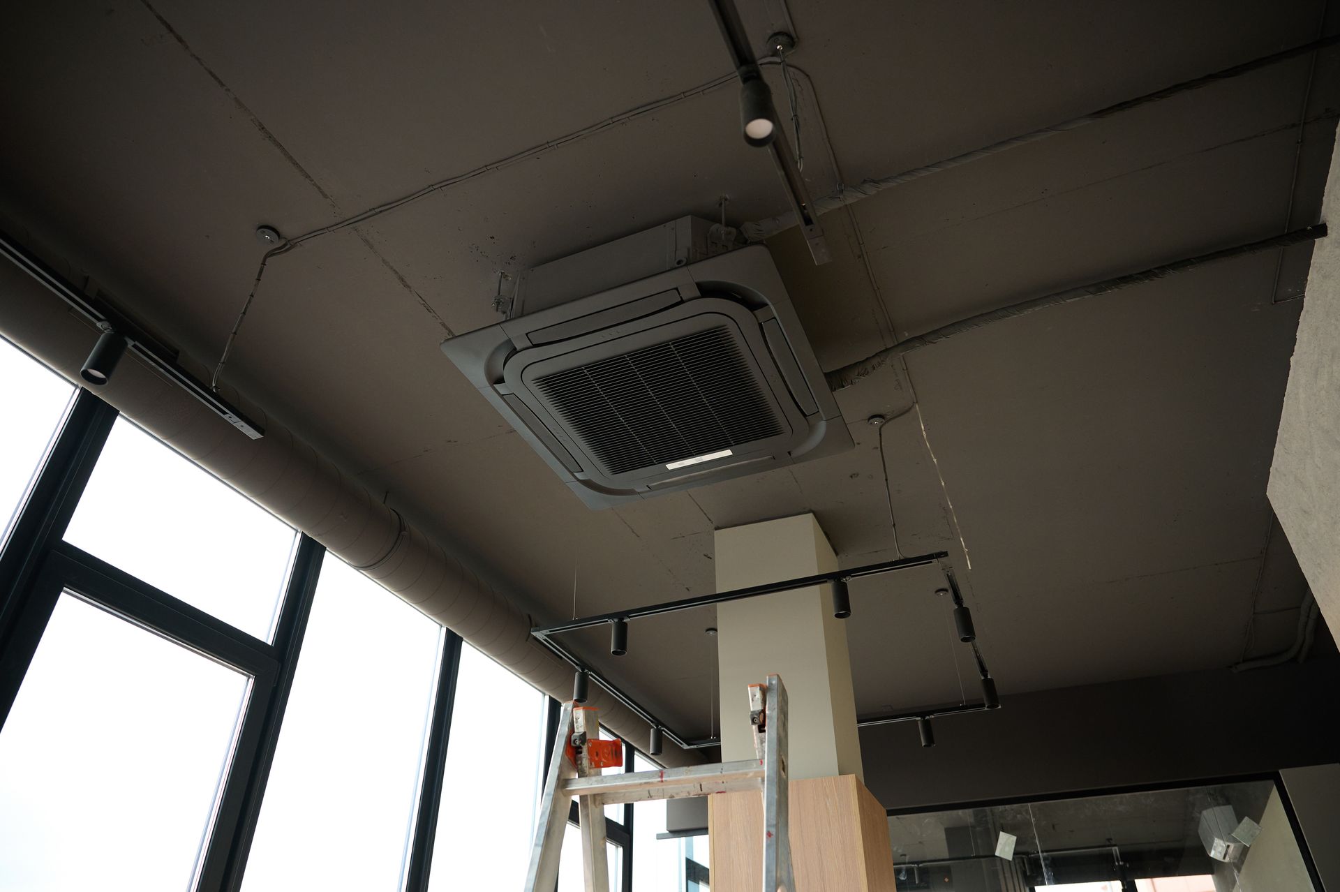 Ceiling-mounted HVAC unit installed in a commercial space with exposed ductwork and ladder below. Ceiling-mounted air vent in a modern interior with windows and hanging cables