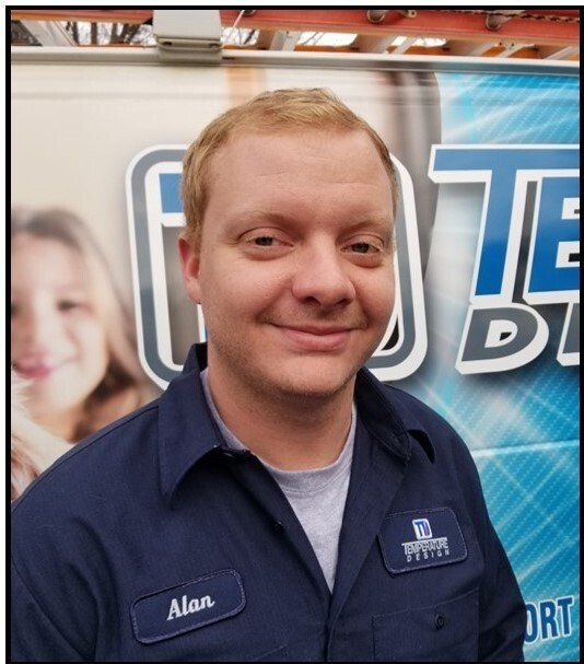 Man in a navy work shirt stands in front of a trade show booth, smiling at the camera.