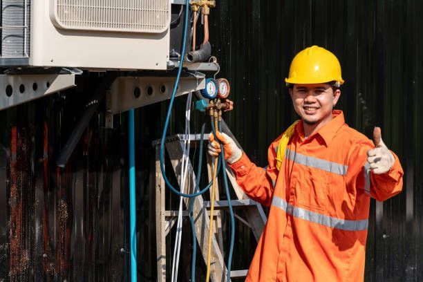 Technician in orange coveralls and yellow hard hat smiling beside electrical equipment, giving thumbs up