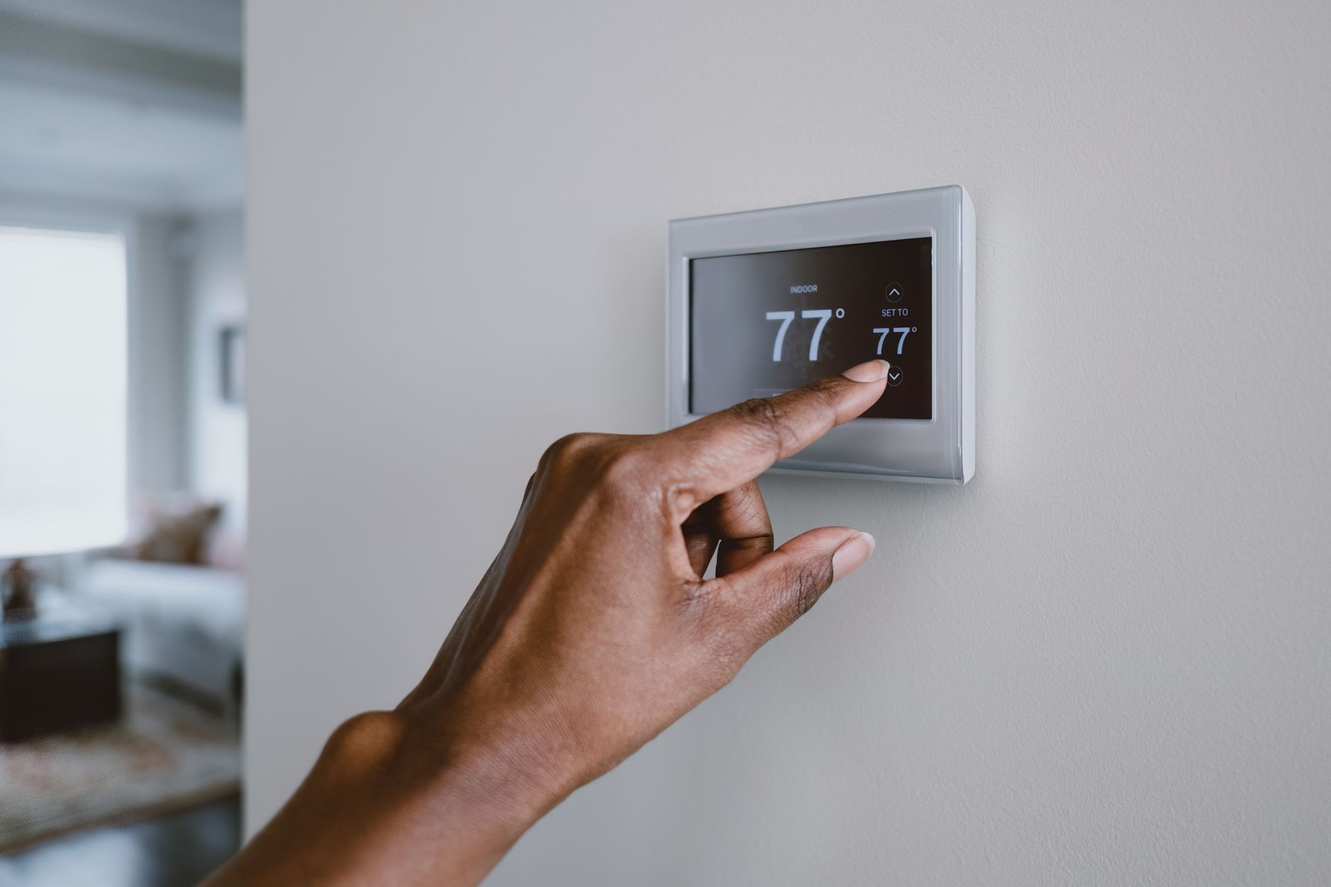 Hand adjusting a wall thermostat displaying 77° in a modern room