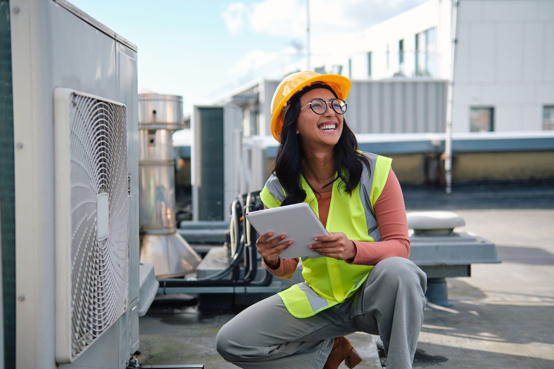 Woman in safety vest and hard hat inspects HVAC unit, using a tablet on a rooftop. Technician in safety vest and hard hat kneels by rooftop HVAC units, holding a clipboard and smiling.