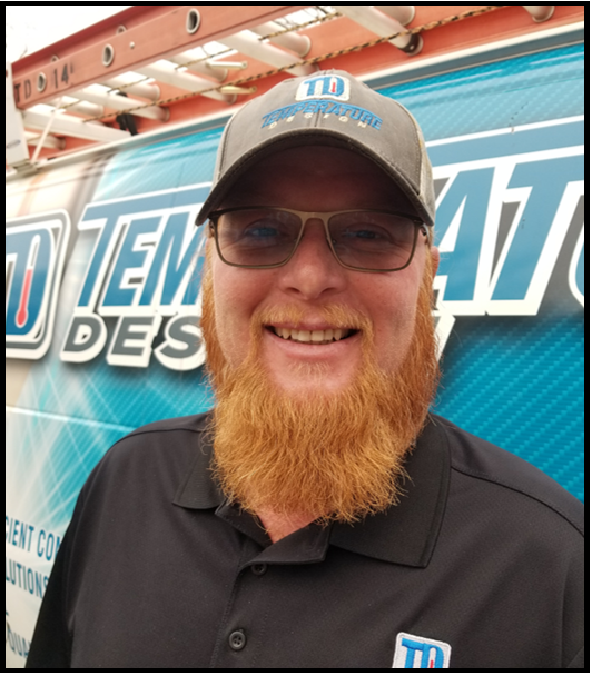 Smiling person with red beard and glasses in a cap and black polo standing before a blue event banner