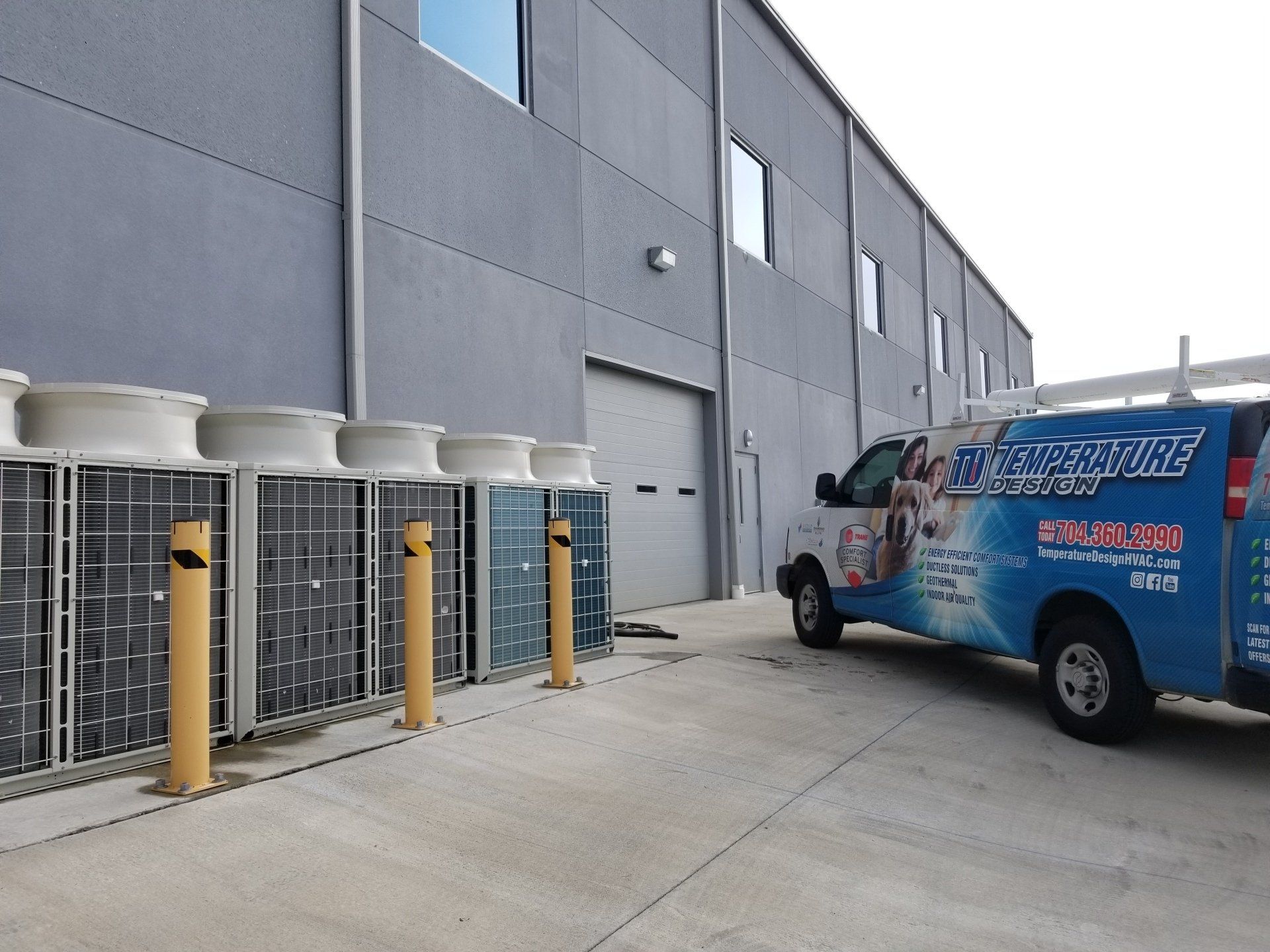Blue service van parked beside an industrial building and HVAC units behind yellow bollards